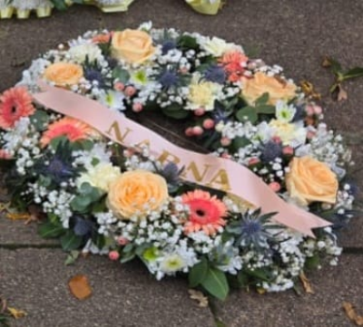 Pastel Memories Wreathe - A delicate pastel wreath created with love and gentle remembrance. This circular tribute features soft cream roses, pink gerberas, lilac blooms, and touches of blue and white seasonal flowers, all arranged with lush foliage for a serene, comforting finish. The wreath includes a personalised tribute ribbon (as pictured with Nana), making it a heartfelt way to honour and celebrate a cherished life.